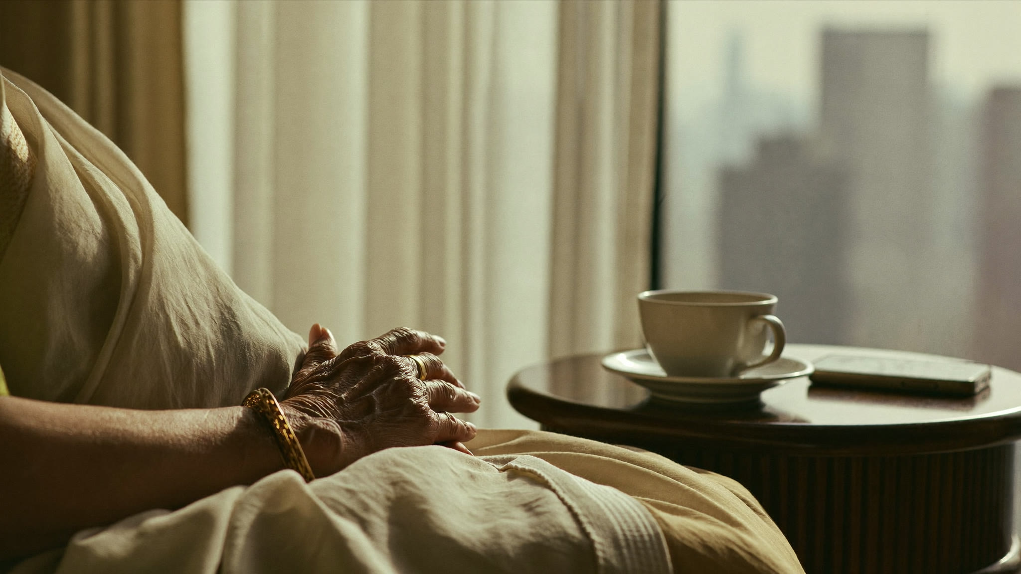 An older woman's hands folded at rest in her lap, a cup of tea and her phone face down on the side table beside her, the city soft and hazy through the curtains behind.